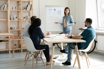 Young Caucasian female speaker or coach make flip chart presentation for diverse colleagues at office meeting. Successful woman trainer present project on whiteboard for employees at briefing.