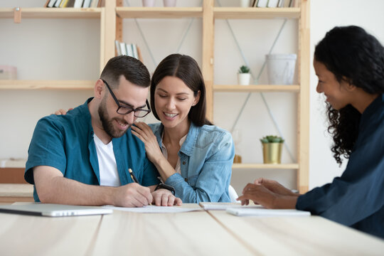 Happy young Caucasian couple put signature on document close deal with female realtor or broker at meeting. Smiling spouses sign paperwork contract make agreement with real estate agent in office.
