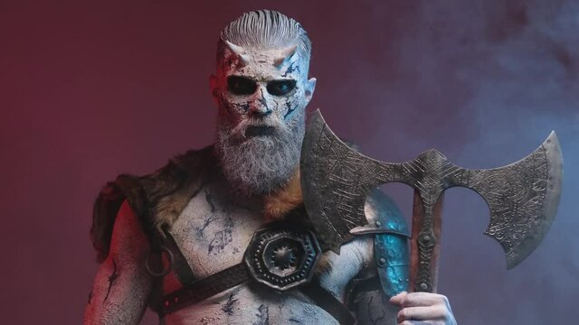 Violent and scary northern undead warrior with pale skin and horns posing in smokey dark red background holdgin two handed double axe and staring at camera with blue eyes.