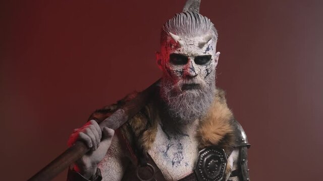 Winter dead warrior with pale skin and beard poses in dark red background with smoke holding his double axe on his shoulder rotating his head and looking around.