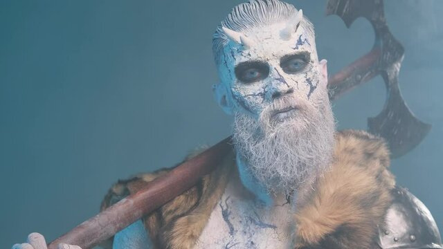 Mystic and dangerous king of the dead with pale skin in light armour with fur posing in dark blue background looking around and holding his double axe on his shoulder.