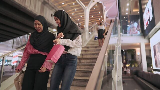 Young multi ethnic female Muslim joyfully talking walking down the shopping mall stair together, non plastics bags concept, downtown shopping on a discount sale, relaxing exercise, wealth cash flow