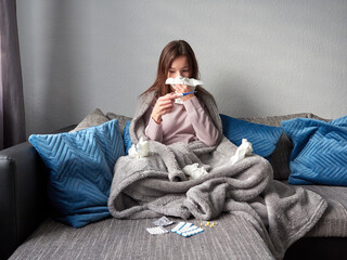 Young sick girl sitting on a couch under a blanket. A woman with a runny nose is blowing her nose...