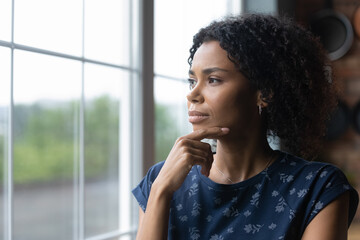 Fototapeta na wymiar Thoughtful millennial african american woman standing near window, recollecting memories or feeling lonely at home. Pensive young biracial lady contemplating, lost in thoughts, vision concept.