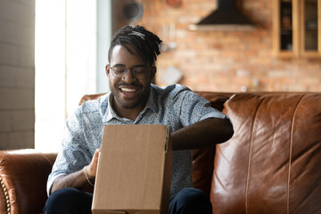 Smiling young african american man in glasses unpacking parcel in carton box, feeling excited of...