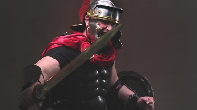 Armed with shield and gladius violent and savage roman warrior in black armour with helmet and red mantle poses in dark background moving from left to right attacking and screaming to camera.