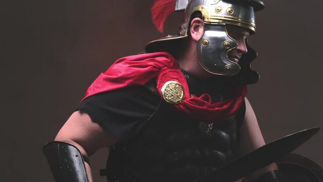 Armed with shield and gladius violent and savage roman warrior in black armour with helmet and red mantle poses in dark background moving from left to right attacking and screaming to camera.