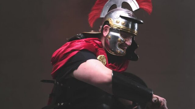 Armed with shield and gladius violent and savage roman warrior in black armour with helmet and red mantle poses in dark background moving and attacks screaming to camera.