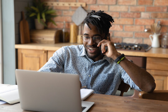 Happy young african american businessman talking on cellphone while working on computer distantly at home. Smiling 30s multiracial man in glasses consulting client by phone call, multitasking concept.