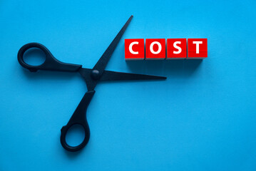 Cut cost concept. Top view of scissors and red wooden block written with COST on a blue background.