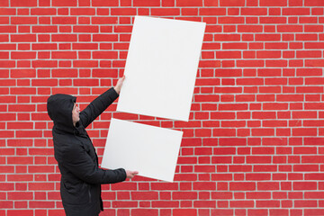 A young man stands against a background of a red brick wall and holds in front of him two white blank canvases for painting