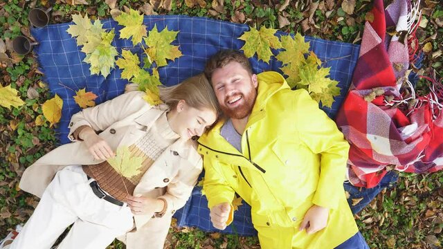 portrait of a happy couple smiling at the camera, lying on a blanket on green grass strewn with yellow leaves in an autumn Park, taken from above.