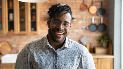 Head shot portrait of happy 30s african american hipster man in eyeglasses, posing in kitchen. Screen view smiling biracial guy holding web camera call conversation pr blogger recording video.