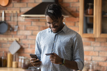 Smiling 30s african ethnicity biracial man in eyeglasses holding plastic bank credit card, checking...