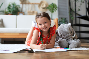 Smilling happy girl lying on warm floor with a toy elephant near to her enjoying creative activity,...