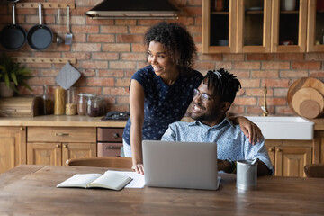 Dreamy happy young african ethnicity married family couple looking in distance, visualizing future or imagining planning holiday vacation, using computer together in kitchen, vision concept.