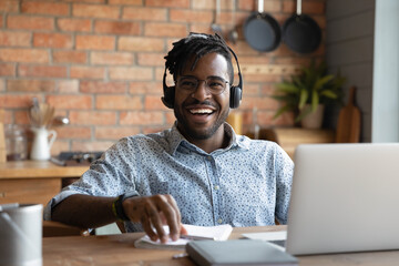 Happy young african american man in eyewear and modern headset sitting at table with computer,...