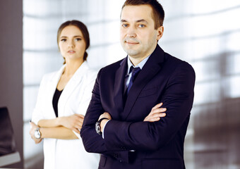 Portrait of a friendly middle aged businessman in a dark blue suit, standing with crossed arms together with a colleague in a modern office. Concept of success in a business
