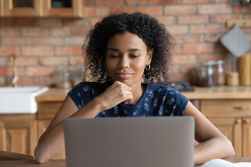 Pleasant thoughtful young african american businesswoman working on computer, sitting at table at home. Happy millennial multiracial woman looking at computer screen, pondering making decision indoors