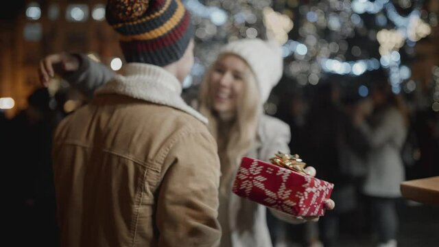 Guy giving present to girl friend on christmas outdoors. Teenagers spending holiday season outside in town, having great time, showing love and friendship.