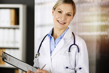 Friendly female doctor standing in sunny clinic. Portrait of cheerful smiling physician. Perfect medical service in hospital