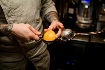 man bartender holds steel squeezer in his hands with slice of orange inside