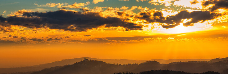 Fototapeta premium Panoramaaufnahme einer Hügellandschaft zur goldenen Stunde mit wolkigem goldenen Himmel