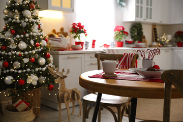 Fototapeta na wymiar Cup of drink and candy canes on wooden table near Christmas tree in kitchen interior