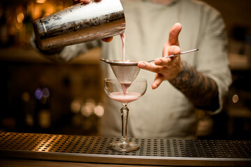 bartender holds sieve over wine glass and gently pours cocktail from shaker.