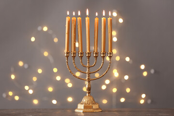 Golden menorah with burning candles on table against grey background and blurred festive lights