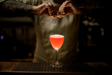 male bartender holding lemon peel over glass with bright cocktail and sprinkles on it.
