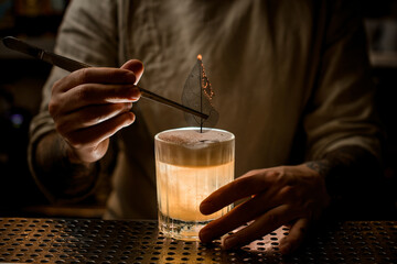 male bartender holds tweezers with smoldering leaf to decorate glass with cocktail