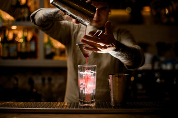 man holds sieve over transparent glass and gently pours cocktail from shaker.