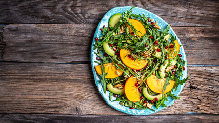 Persimmon, avocado salad with pumpkin seeds, walnuts, pomegranate and arugula on a wooden background. Long banner format. space for text top view