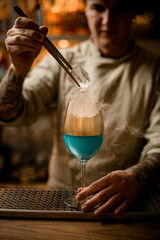 male barman holds piece of ice over steaming wine glass decorated with gold