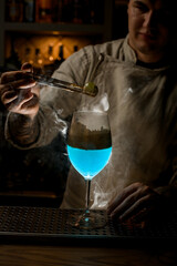 bartender at dark bar holds golden piece of ice over steaming wine glass and look at it