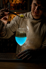 young bartender at dark bar holds golden piece of ice over steaming wine glass and look at it