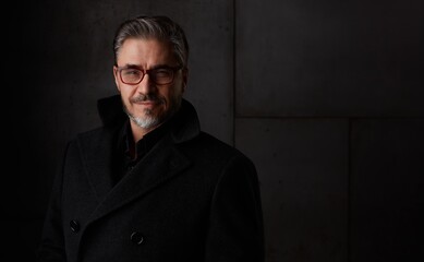 Portrait of happy older man in 50s with gray hair and glasses against dark background. Wearing coat, smiling, copy space.