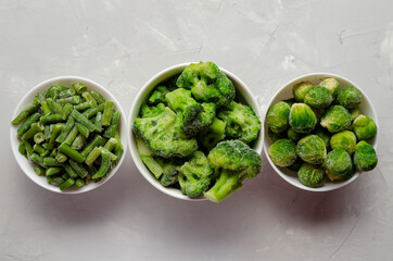 A mix of frozen green vegetables: beans peas broccoli Brussels sprouts in bowls on a grey background.