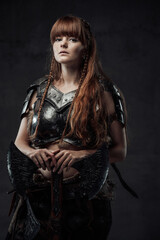 Naklejka na ściany i meble Beautiful but dangerous warlike woman viking with long brown hairs in dark armour holding huge axe in dark background.