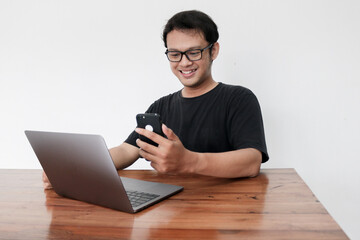 Young Asian man feeling happy and smile what he see in the smartphone and laptop on the table. Indonesia Man wear black shirt Isolated grey background.