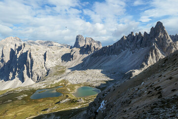 Small, navy blue lakes at the bottom of the valley in Italian Alps. The lakes are surrounded by high and steep peaks. The slopes are lush green. The sky is full of soft clouds. Raw landscape. Remedy