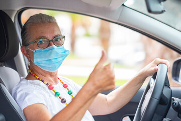 Naklejka premium Coronavirus. An attractive elderly woman inside her car is wearing a surgical mask due to the coronavirus, looks at the camera with a raised thumb