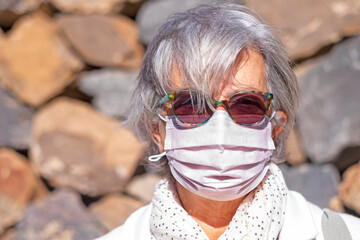 Coronavirus. Front view of a grey haired senior woman standing outdoor in a windy day looking at camera. Wearing surgical mask due to coronavirus infection