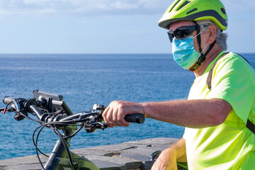 Naklejka premium Coronavirus. Portrait of a senior white haired man with bicycle and helmet doing sport activity enjoying nature and sea, wearing a protective face mask due to coronavirus contagion