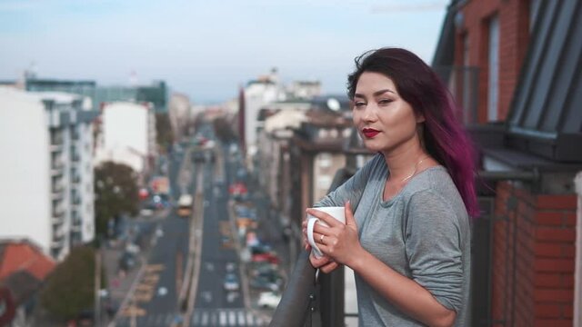 Girl watching a beautiful urban view and street and relaxing alone. Pretty young woman in stays on balcony holds cup of hot coffee or tea.