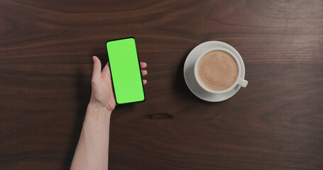 Top view man drinking hot chocolate and using smartphone with green screen over black walnut wood table