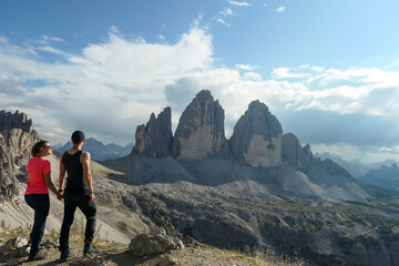 Obraz premium A couple in hiking outfit holding hands on top of Sextner Stein and enjoying the view on the famous Tre Cime (Drei Zinnen) in Italian Dolomites. Desolated and raw landscape, full of lose stones. Love