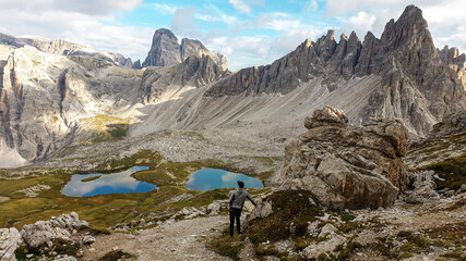 A man hiking with the view on small, navy blue lakes at the bottom of the valley in Italian Alps. The lakes are surrounded by high and steep peaks The sky is full of soft clouds. Raw landscape. Remedy