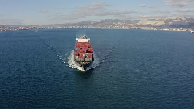 Aerial front view of ultra large red cargo ship with containers floating on the sea waves on warm sunny day. Concept of work on the delivery of sea goods and cargo, drone footage.